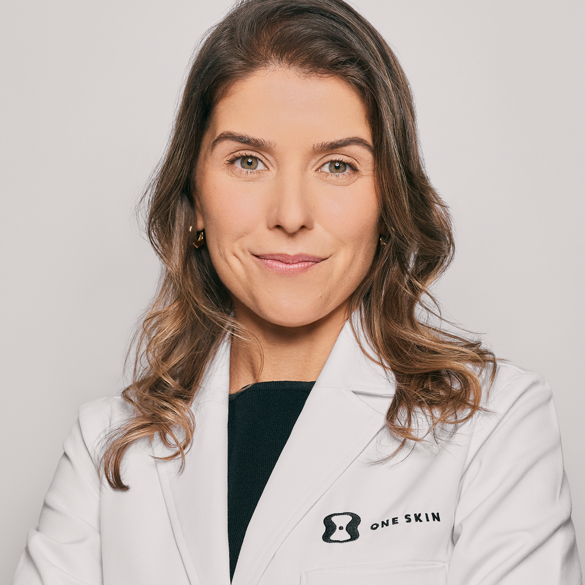 A professional portrait of a woman with brown hair wearing a white lab coat with the 'ONE SKIN' logo on the left chest, set against a solid light gray background.